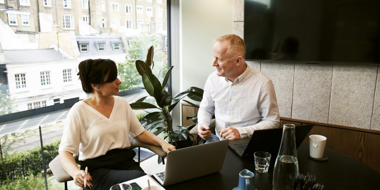 two people sit at a desk facing each other in a sun-lit office with a computer monitor in front of them a TV on the wall behind them.