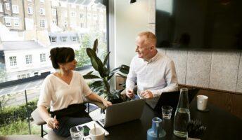 two people sit at a desk facing each other in a sun-lit office with a computer monitor in front of them a TV on the wall behind them.
