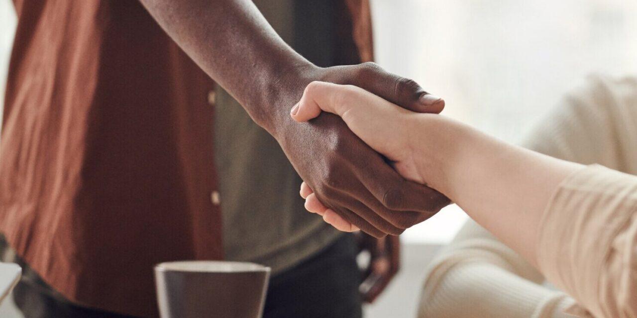 two people shake hands across a conference table where one person is standing and one person is seated
