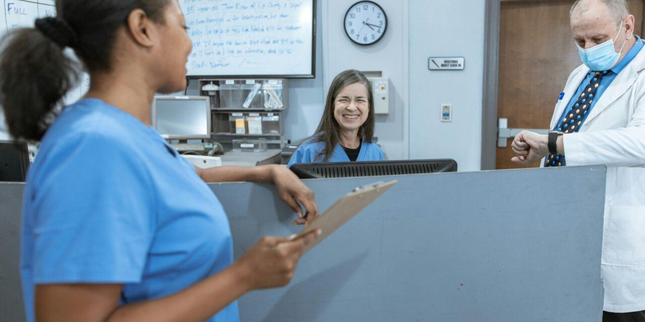 Two nurses in blue scrubs smile at each other over a desk in a healthcare setting while a doctor checks his watch