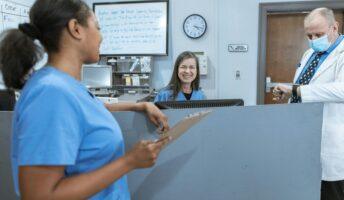 Two nurses in blue scrubs smile at each other over a desk in a healthcare setting while a doctor checks his watch