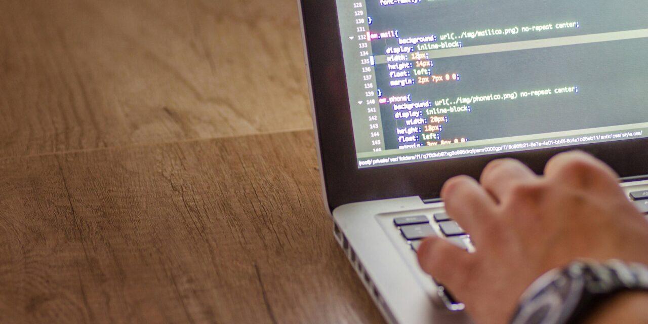 A hand rests on a silver laptop and types computer code which is visible on the laptop screen