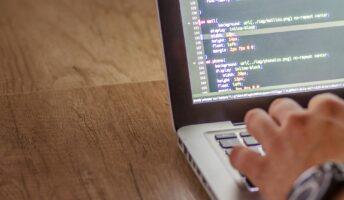 A hand rests on a silver laptop and types computer code which is visible on the laptop screen