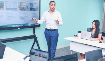 a team in business casual clothing sits at a conference room looking at their team leader presenting content on a large screen