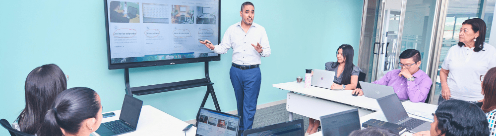 a team in business casual clothing sits at a conference room looking at their team leader presenting content on a large screen