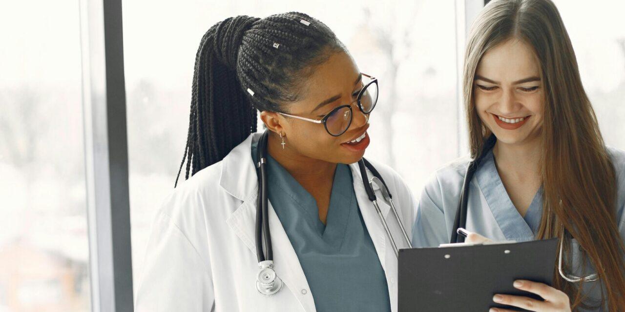 A doctor in a white medical coat and a nurse in blue scrubs look at a clipboard.