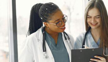 A doctor in a white medical coat and a nurse in blue scrubs look at a clipboard.