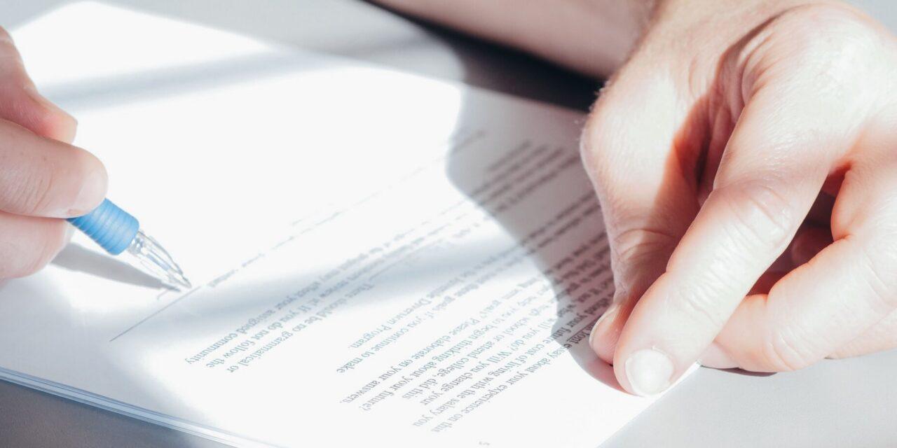 Two hands rest on a table near a paper contract which is about to be signed