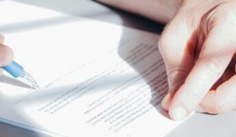 Two hands rest on a table near a paper contract which is about to be signed