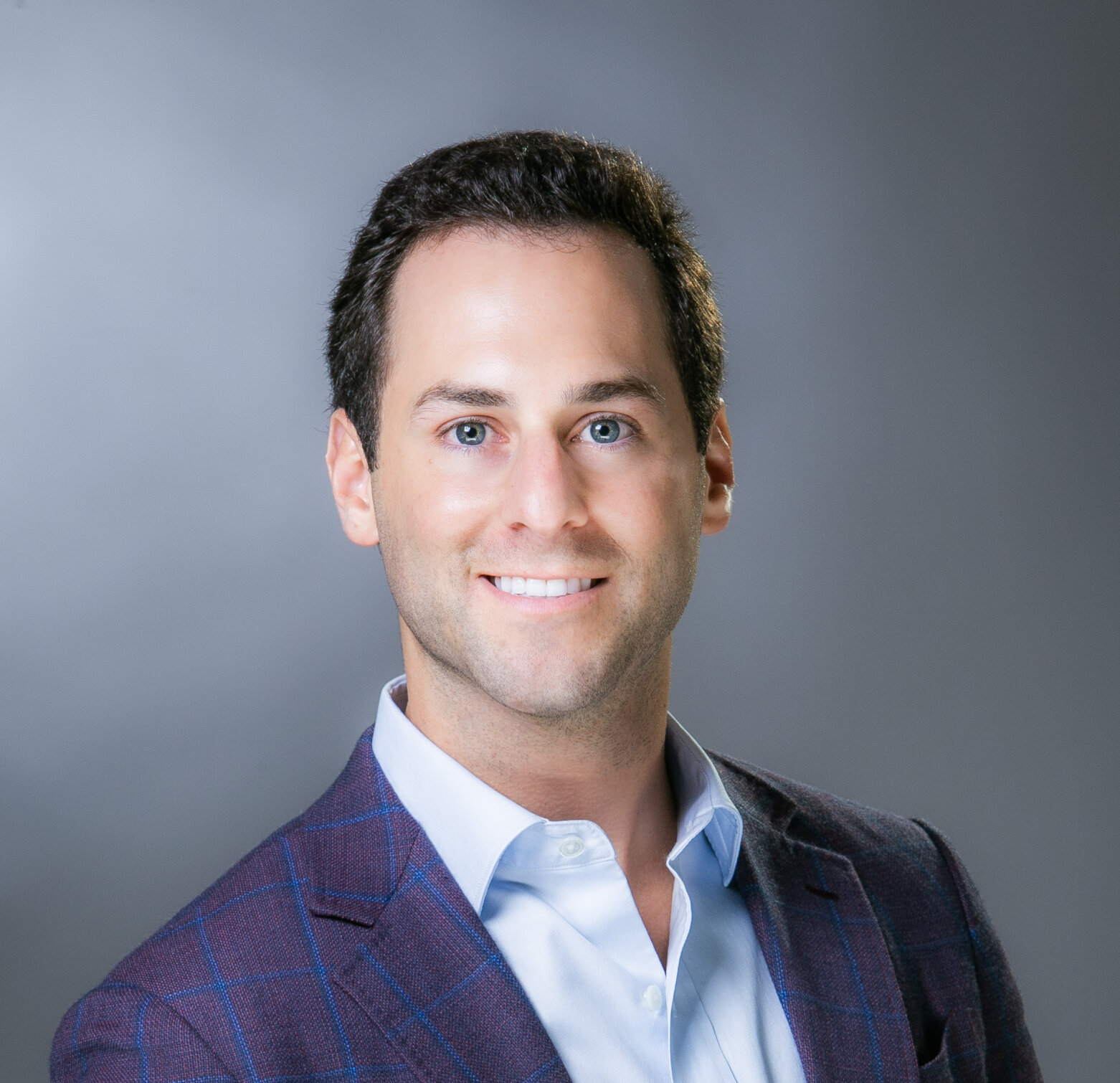 Portrait of a smiling man with dark hair in a plaid blazer against a gray backdrop