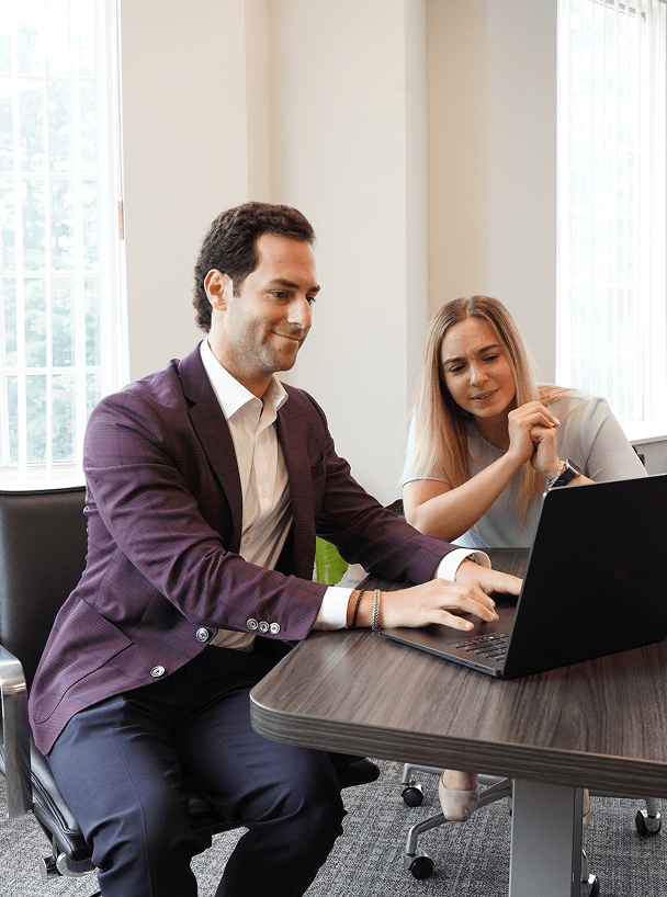 Smiling man in a purple blazer working on a laptop next to a woman in an office meeting room